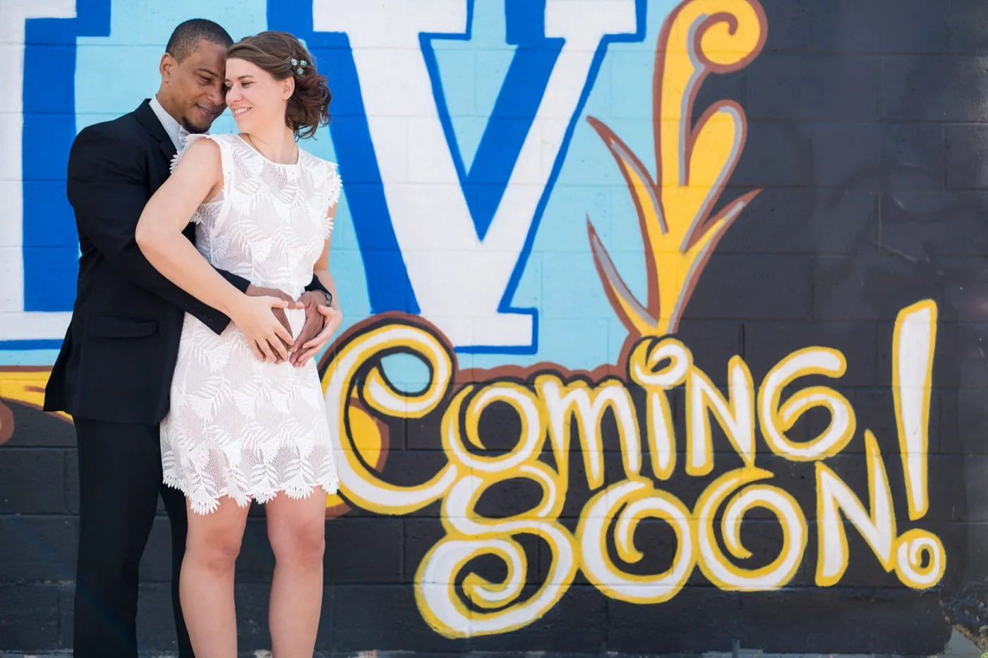 Groom in a black suit with his cheek against his bride's temple in front of a blue LV mural and yellow Coming Soon script