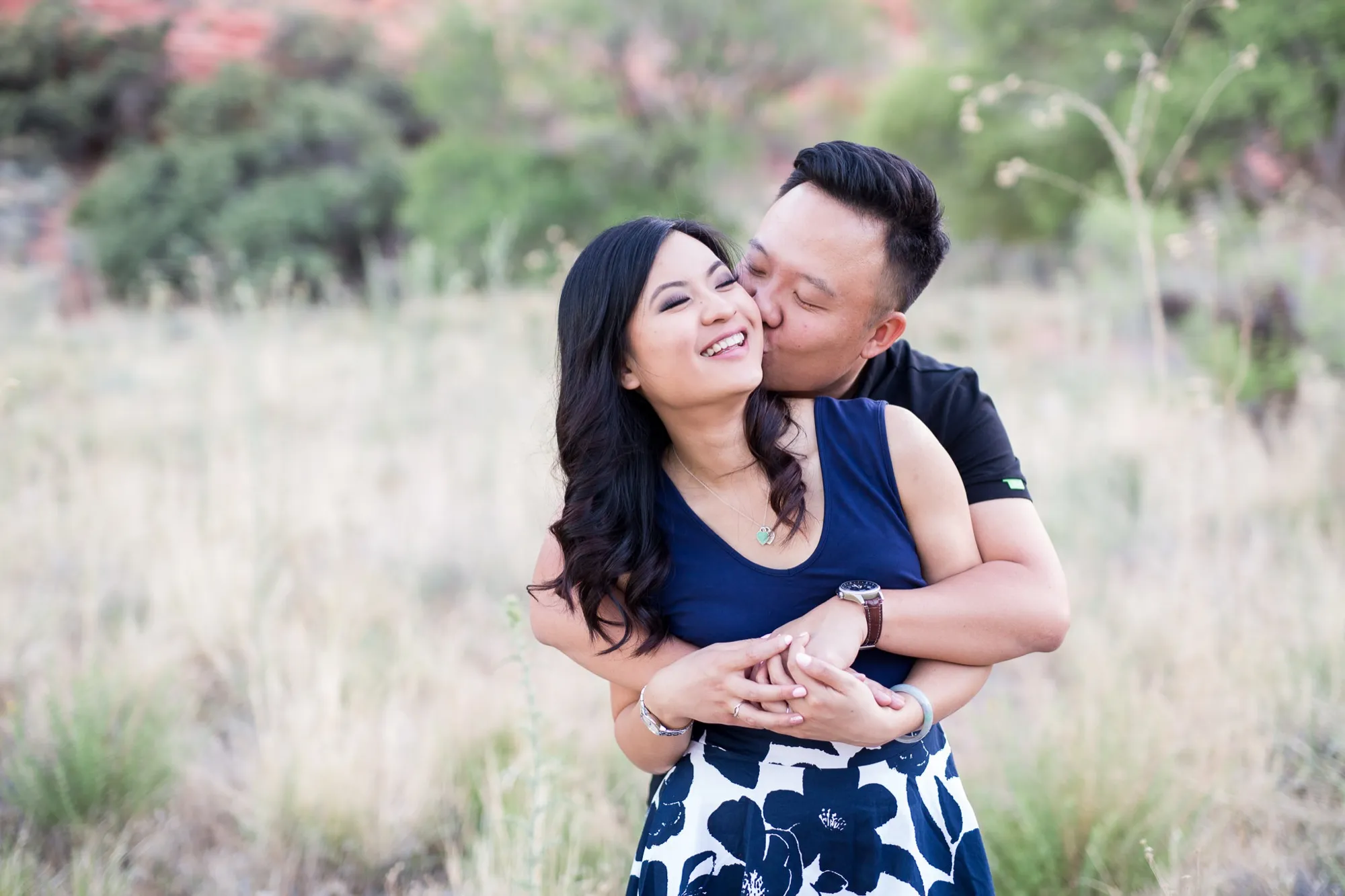A groom-to-be kissing his fiancée on the cheek while she laughs during a desert engagement session outside Las Vegas