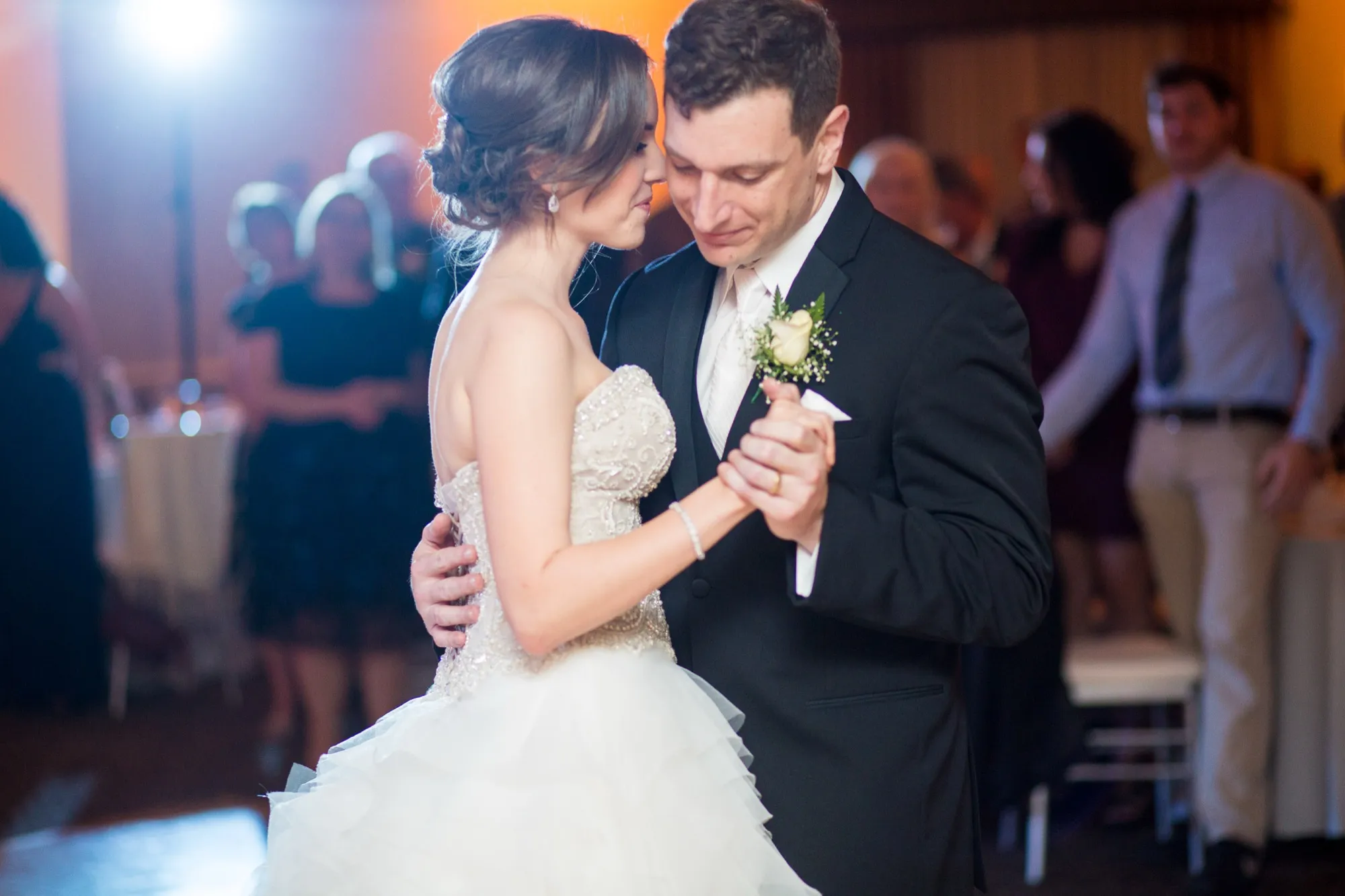 Bride in a beaded gown and groom in a black tuxedo dance close in a dark reception room, guests behind them