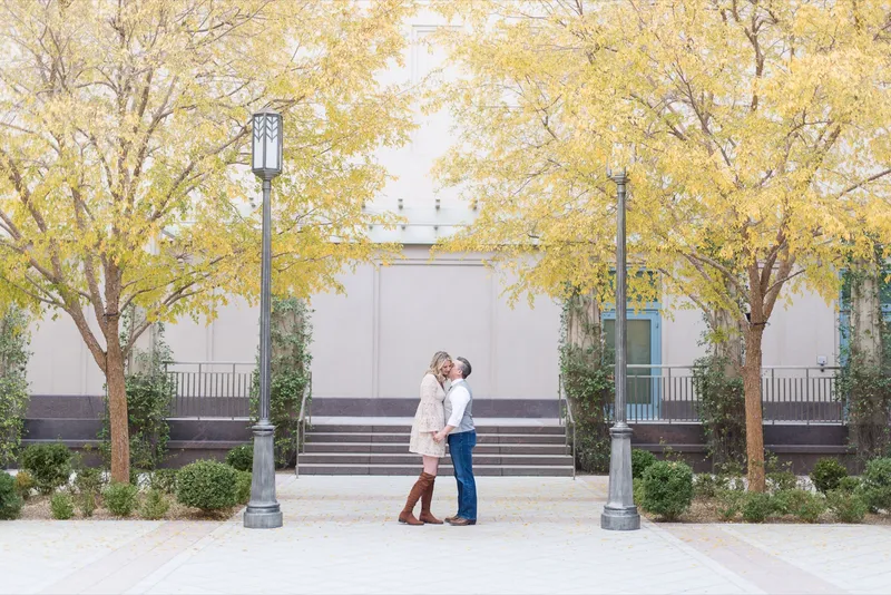 Couple standing close together on a wide stone walkway under two trees in full yellow autumn leaf, fallen leaves scattered on the pavement