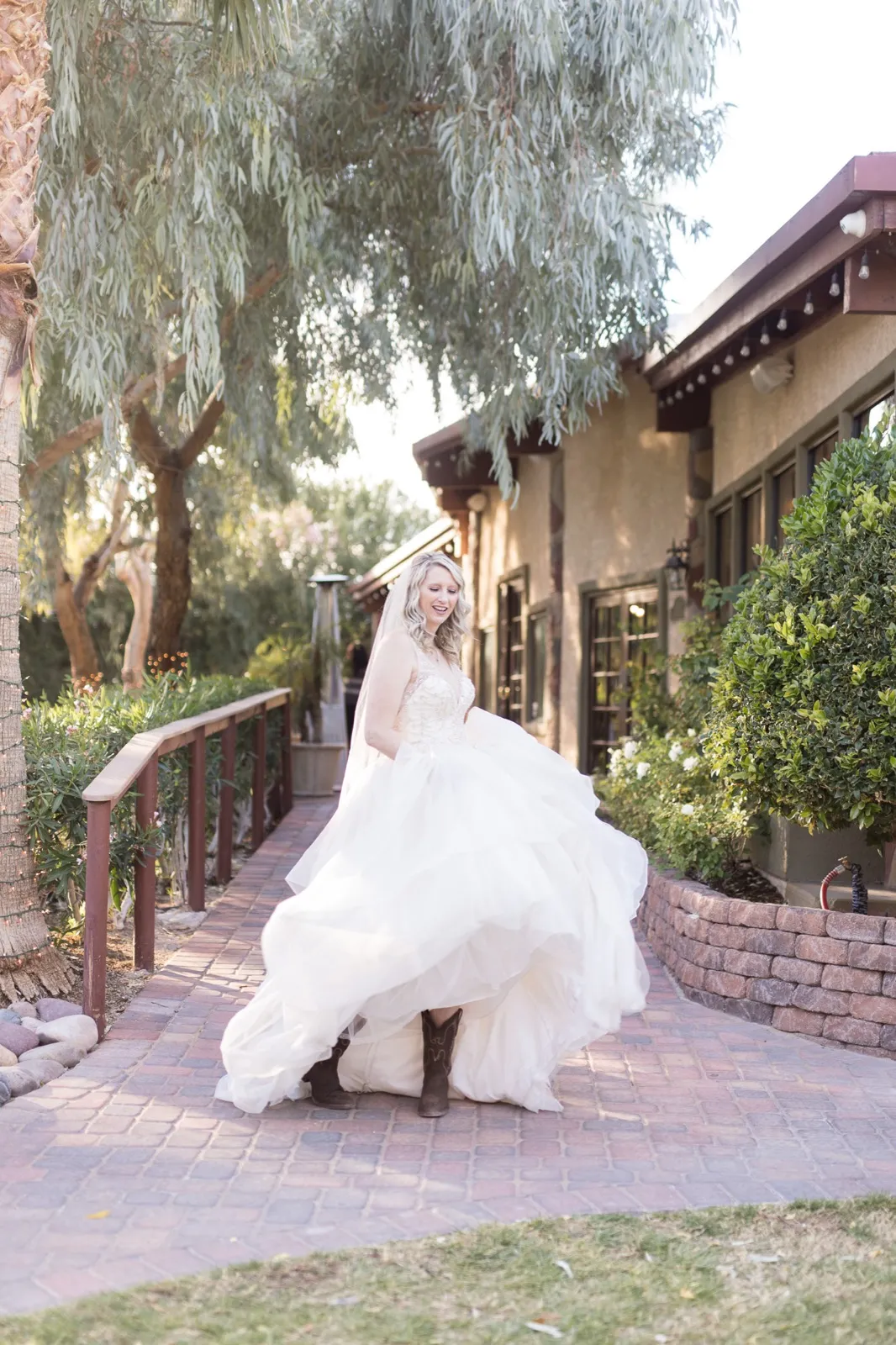 A bride in cowboy boots twirling her tulle gown on a brick path beside a tan stucco building
