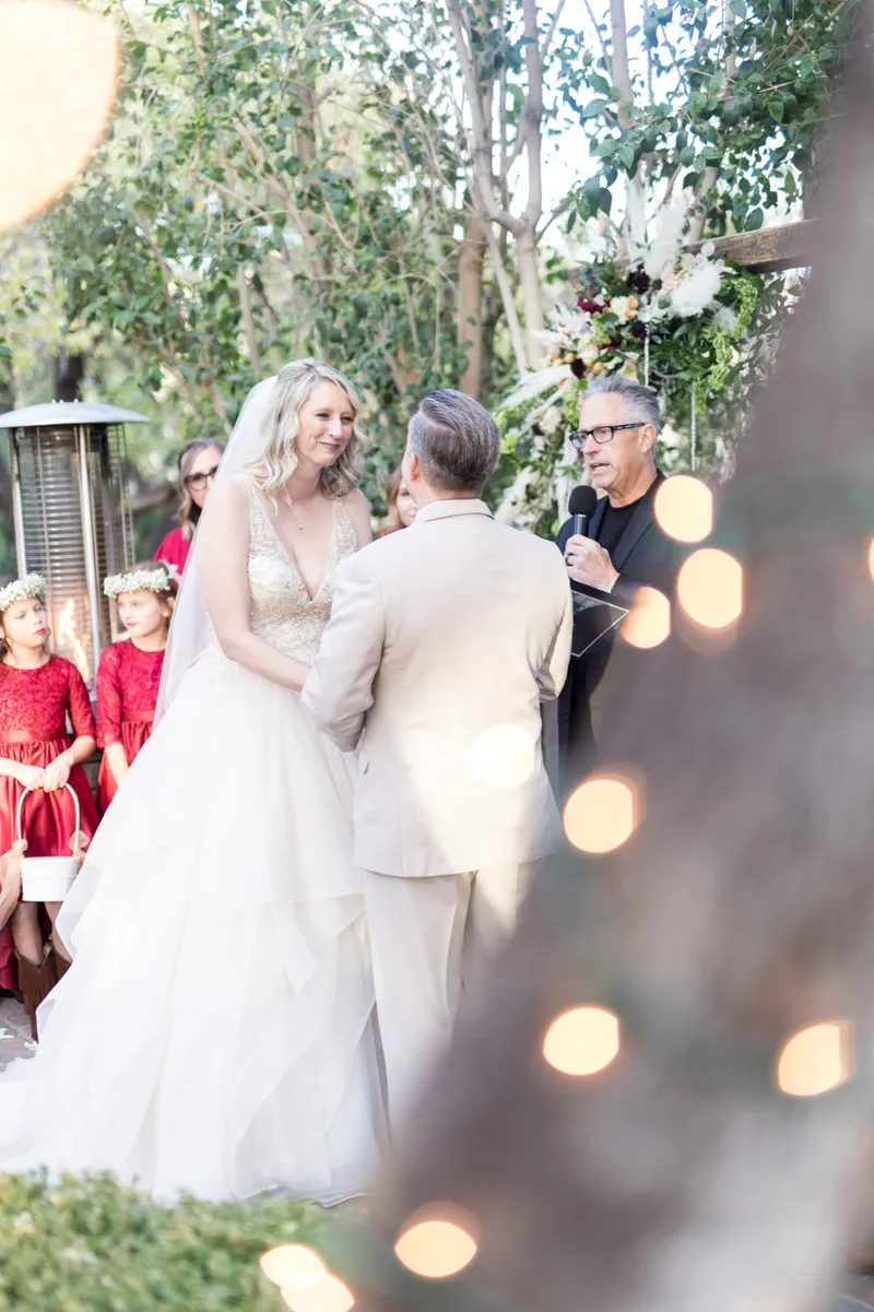 A bride in a ruffled gown and a groom in a tan suit facing each other at the floral arbor with flower girls in red beside them