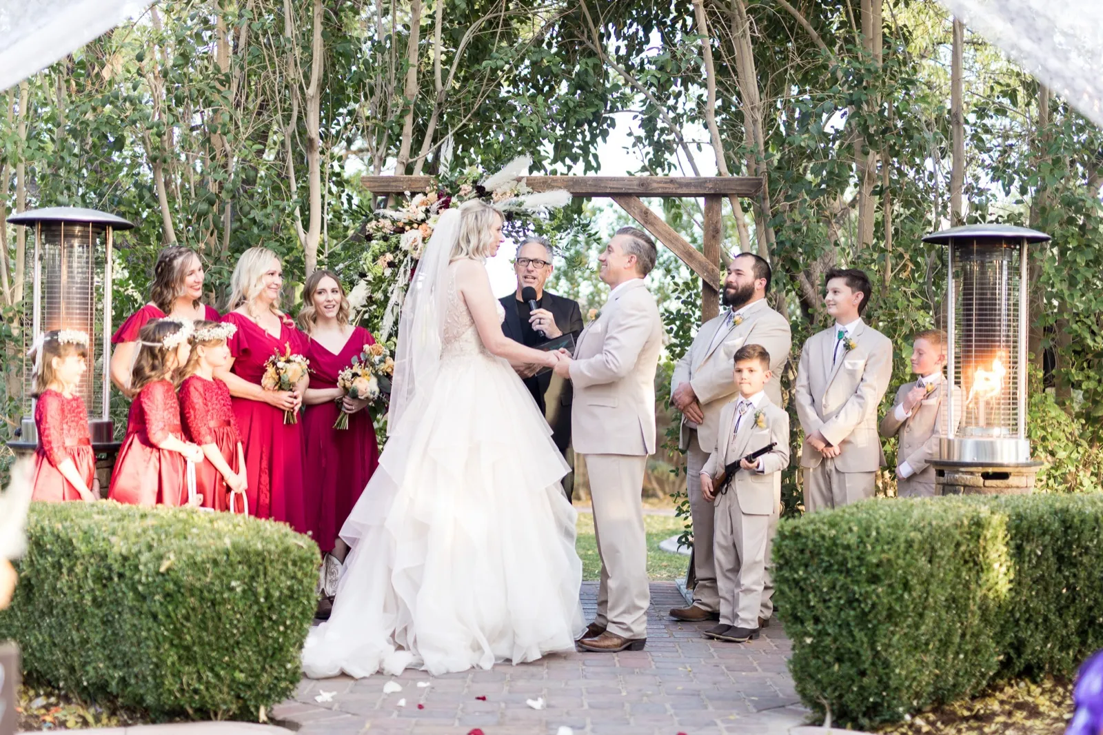 A couple holding hands at a wooden floral arbor, wedding party in red and tan beside tall patio heaters
