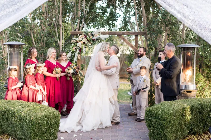 The couple's first kiss under the floral arbor while the wedding party and the flower girls watch