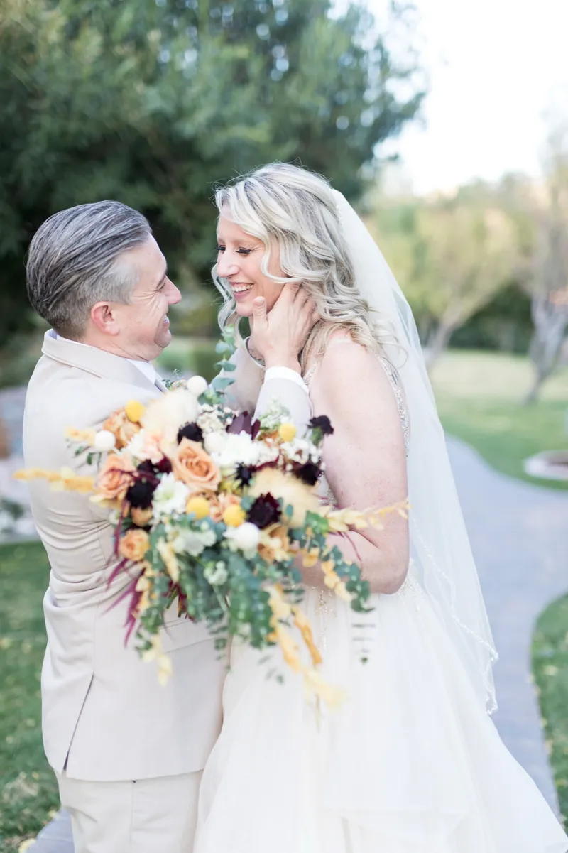 The bride touching the groom's face as they smile at each other, her orange and burgundy bouquet held between them