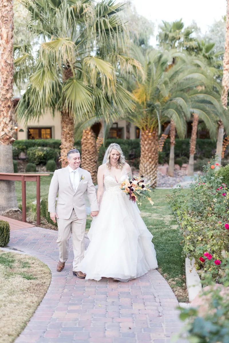 The bride and groom walking a brick garden path lined with palms, clipped hedges and flower beds