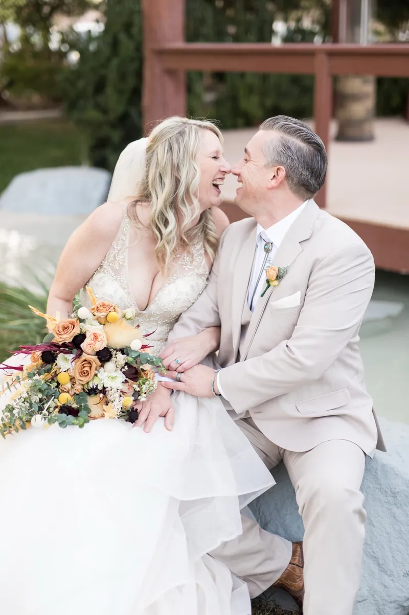 The couple laughing nose to nose while sitting on a boulder at the edge of the pond with the wooden deck behind them