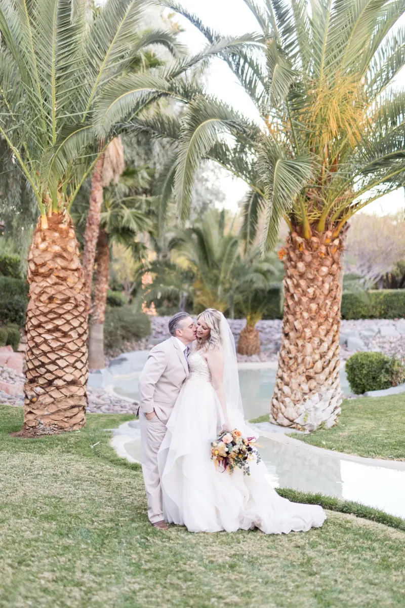 The couple kissing on the grass between two tall palm trunks in late afternoon light