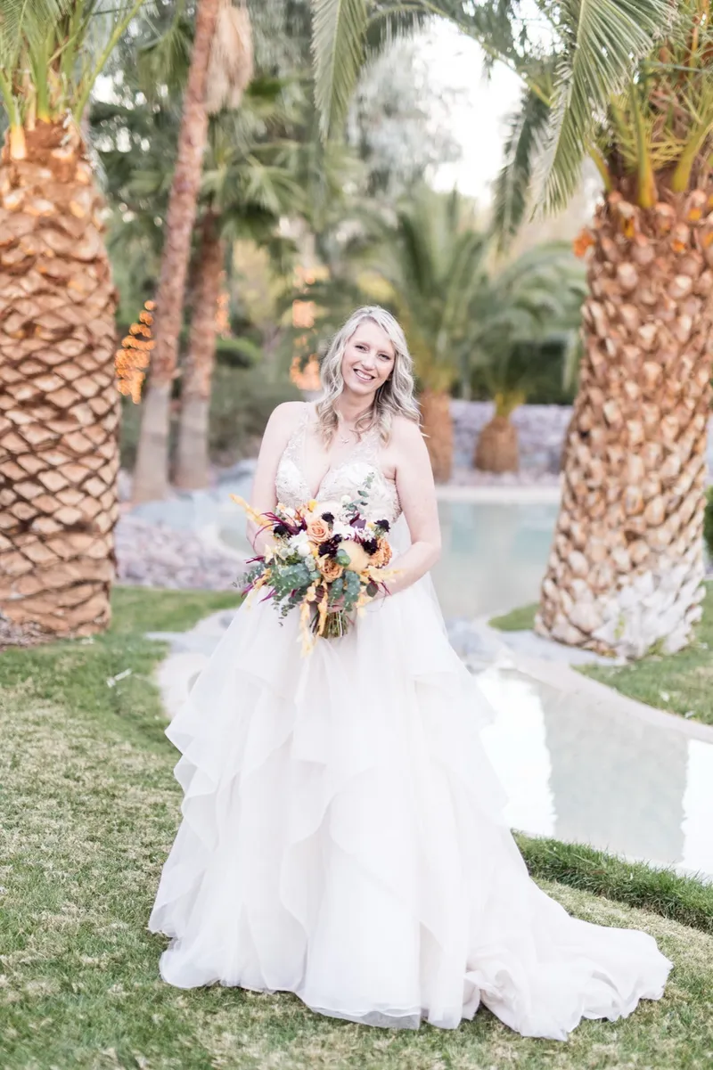 The bride standing alone on the lawn between the palms holding her orange and burgundy bouquet