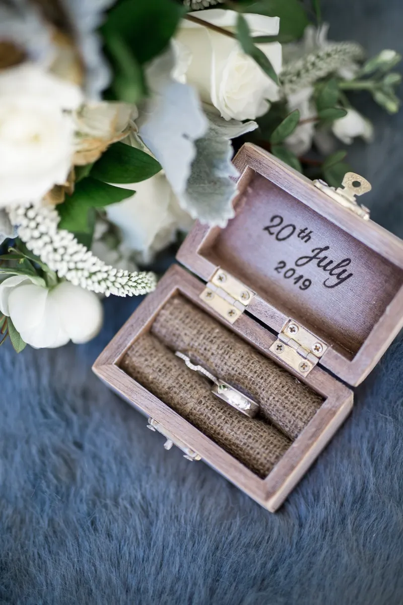 Open wooden ring box holding both bands, the lid printed with the wedding date, set beside a white bouquet
