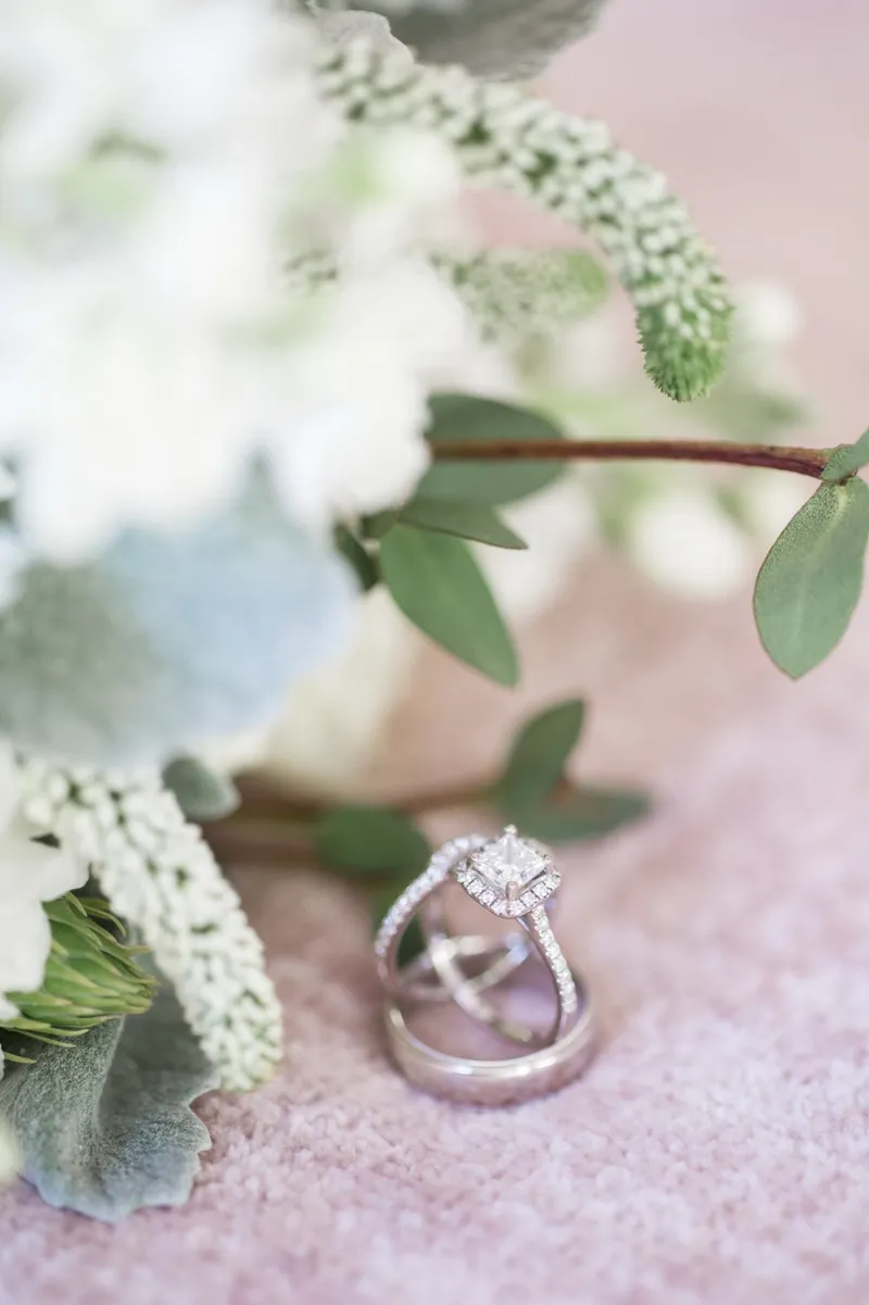 Engagement ring and two bands tucked into blush fabric beside white flowers and grey green leaves