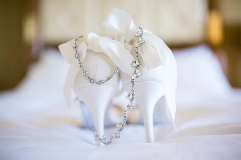The bride's white heels standing on the hotel bedding with a crystal necklace draped between them