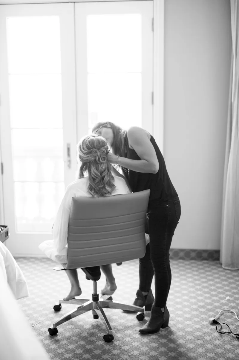 Black and white photo of a stylist leaning in to finish the bride's hair in the light of the hotel room window