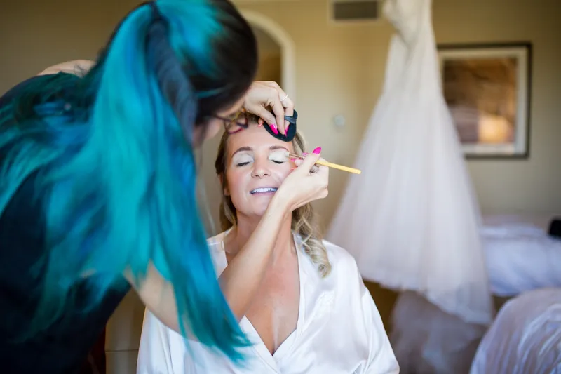Makeup artist with bright teal hair working on the bride's eyes while she sits in her robe, gown hanging behind them