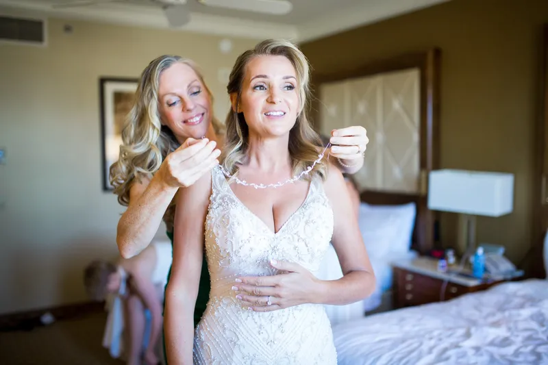 Two women laughing together as one fastens a pearl necklace at the bride's collar