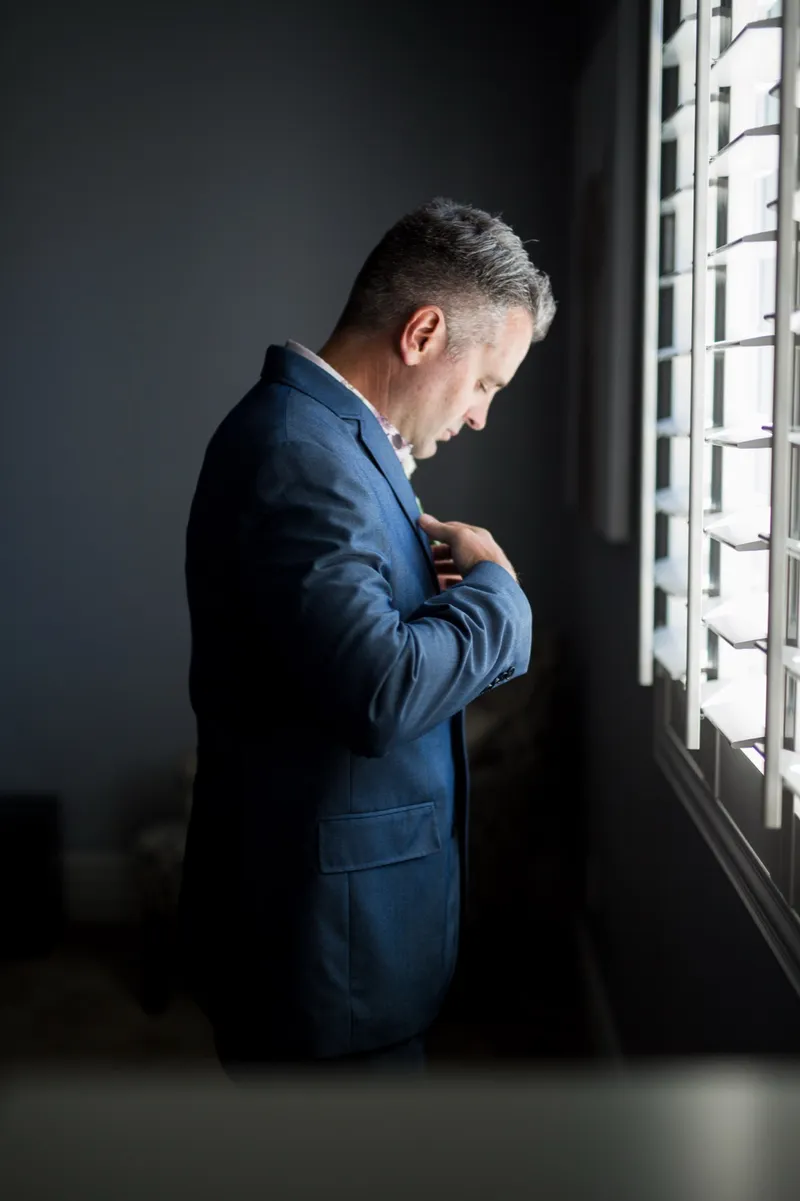 Groom in a blue suit buttoning his jacket beside a bright window, the wall behind him dark