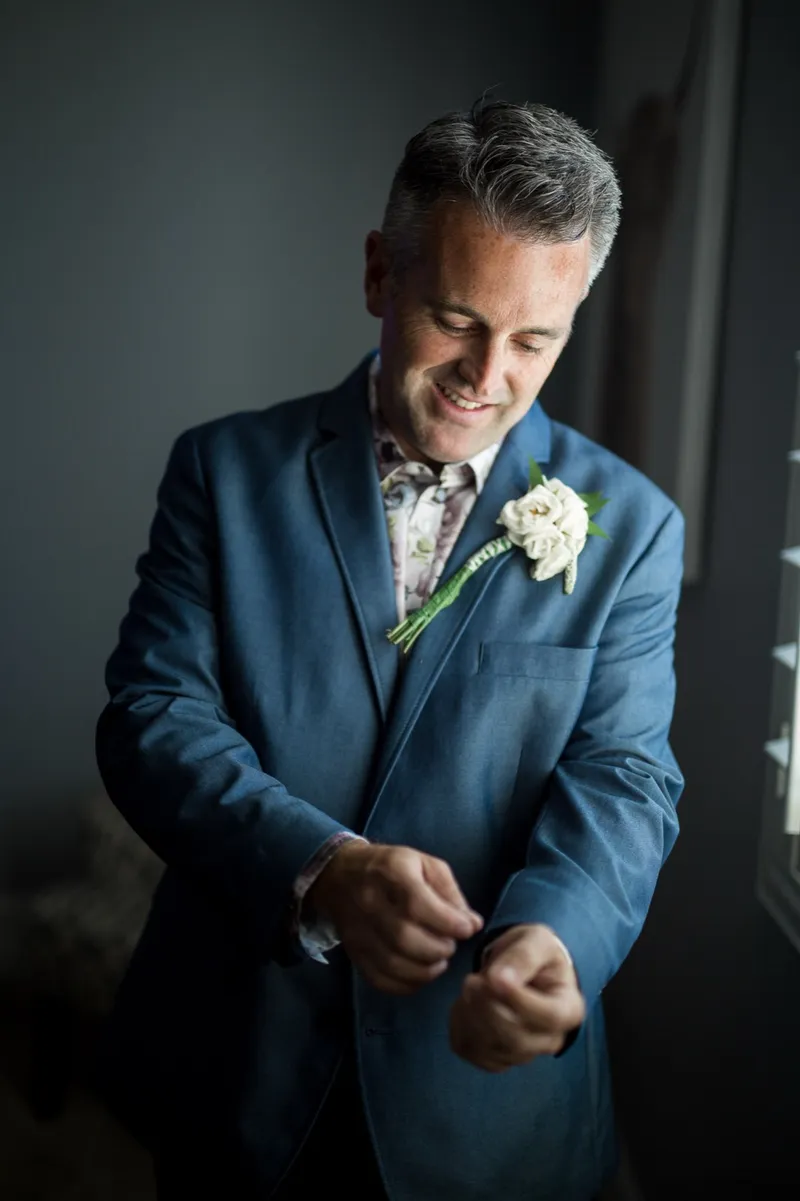 Groom smiling down at his cuff as he fastens it, a white boutonniere already pinned to his lapel
