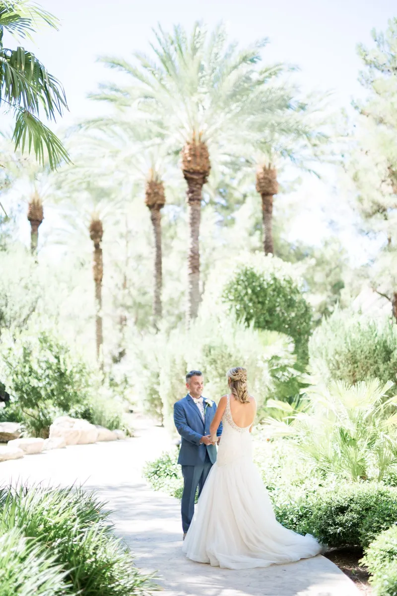 Bride and groom holding each other on a wide paved garden path with clipped hedges and tall palms behind them