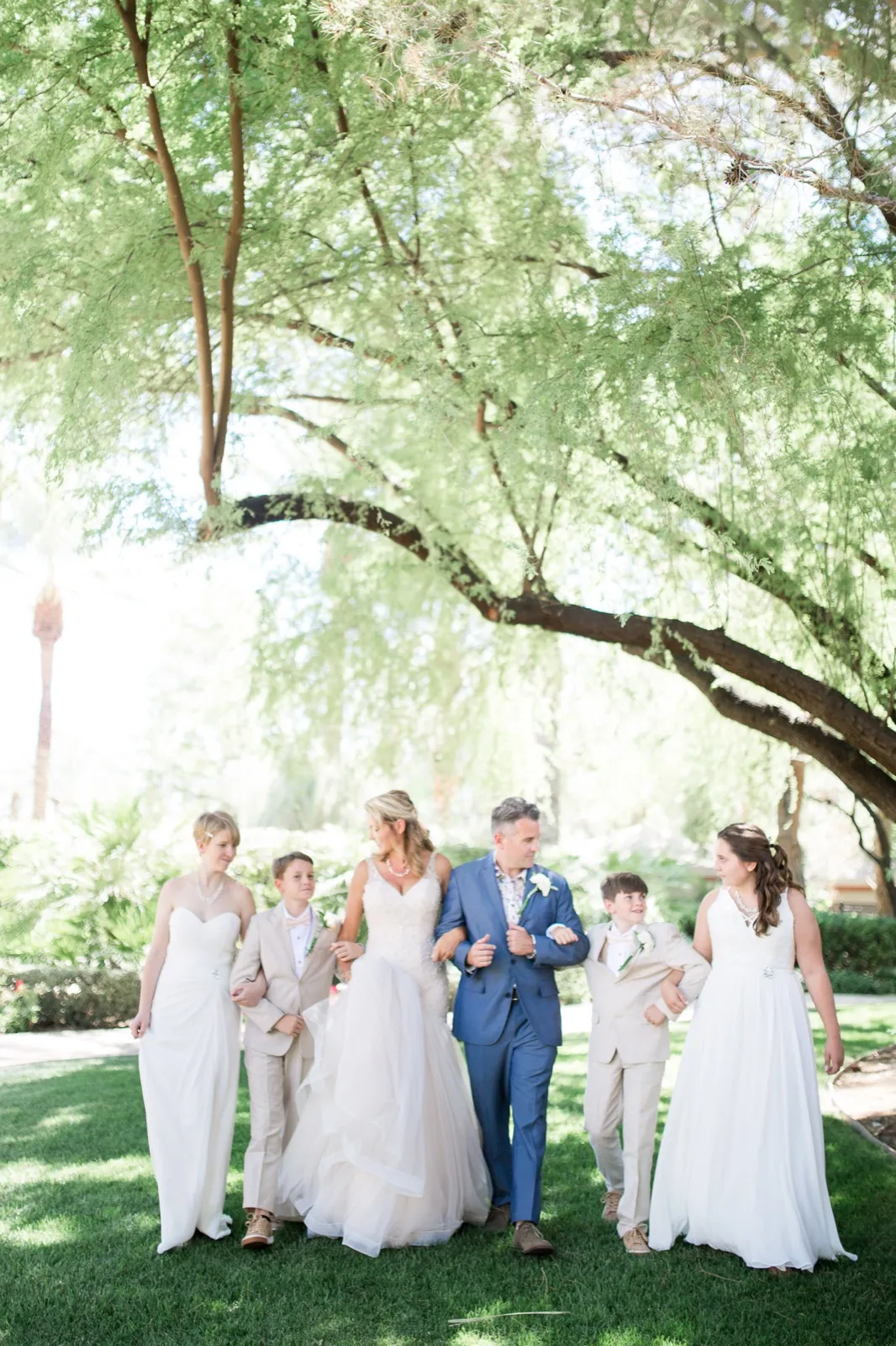 Bride and groom walk arm in arm with four children in ivory dresses and tan suits under a wide green tree