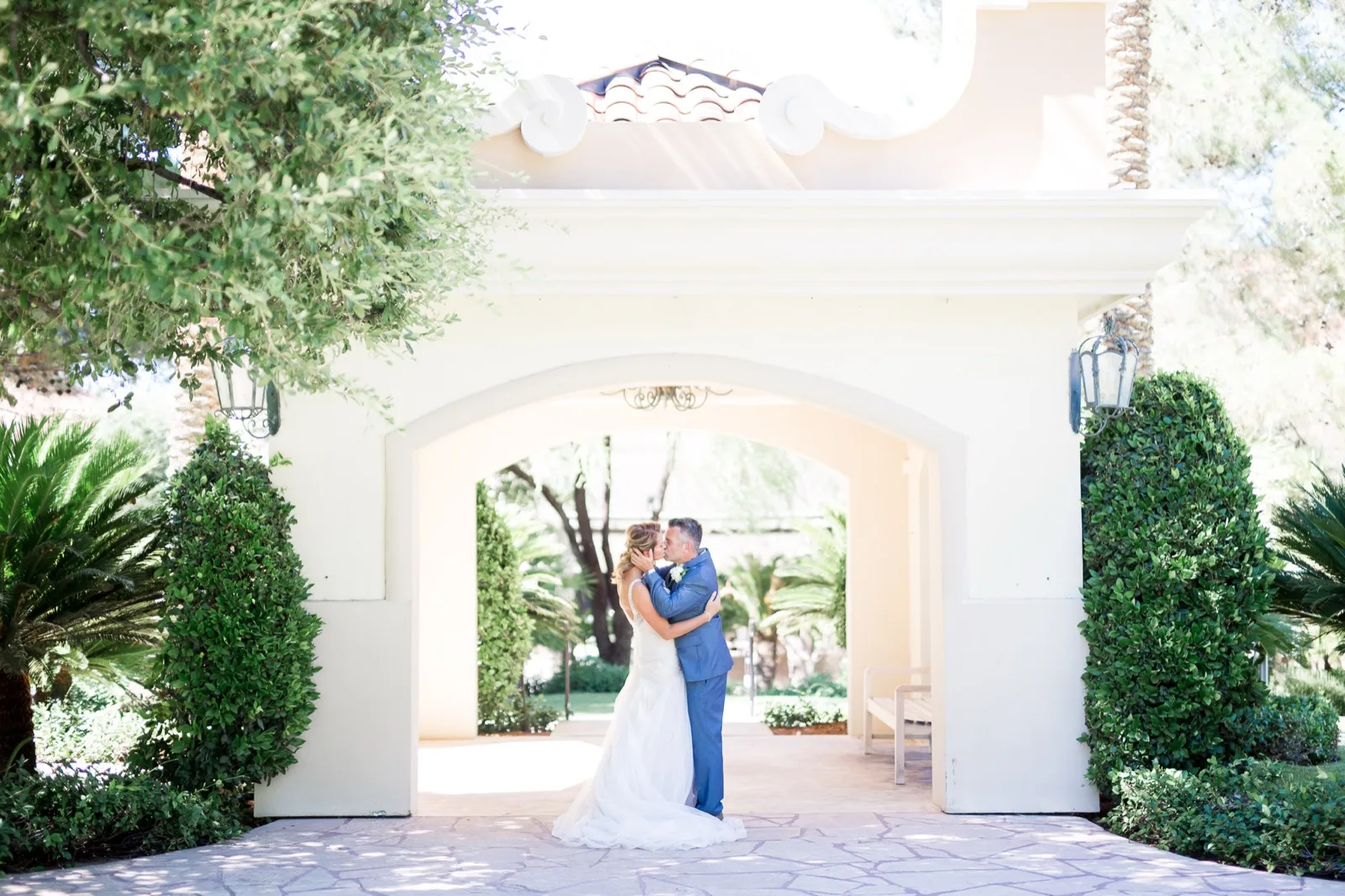 Bride and groom kissing under a wide stone archway with tall hedges on either side and palms in the garden behind them