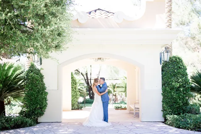 Bride and groom kissing under a wide stone archway with tall hedges on either side and palms in the garden behind them