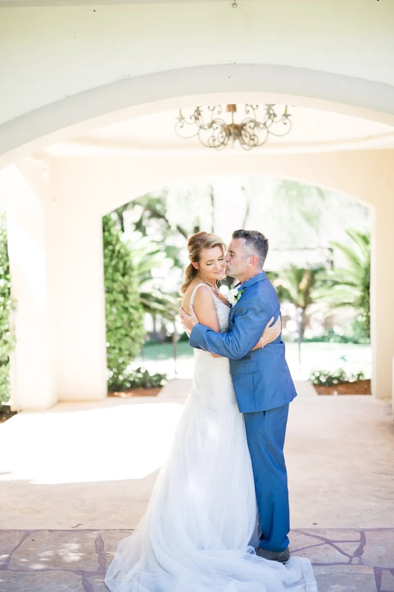 Bride and groom holding each other under the archway with a chandelier hanging above them and green garden showing through the far arch