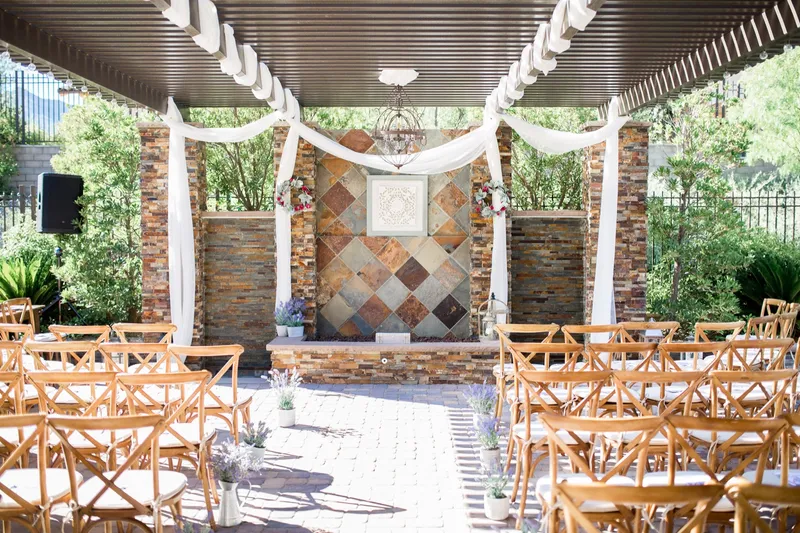 Backyard ceremony set under a slatted pergola with white drapes, wooden cross back chairs in rows, potted lavender along the aisle and a speaker at the side