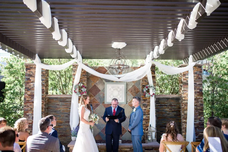 Bride and groom standing with the officiant under the draped pergola, guests seated close on either side