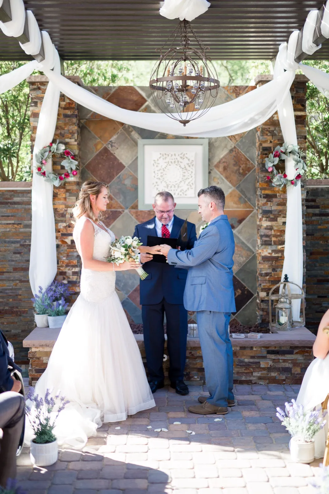 Officiant in a navy suit holds an open book between a bride and groom exchanging rings at a tiled pergola altar