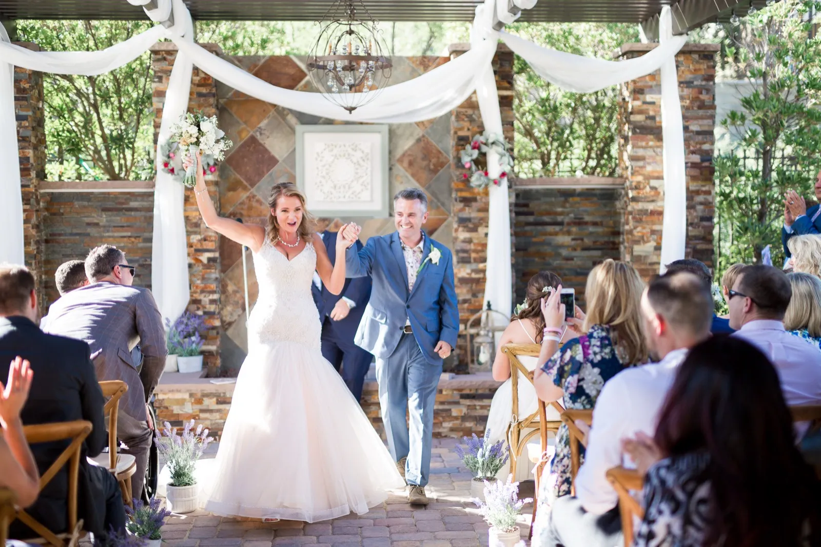 A bride raises her bouquet as she and her husband walk back down the aisle past applauding guests under a stone pergola