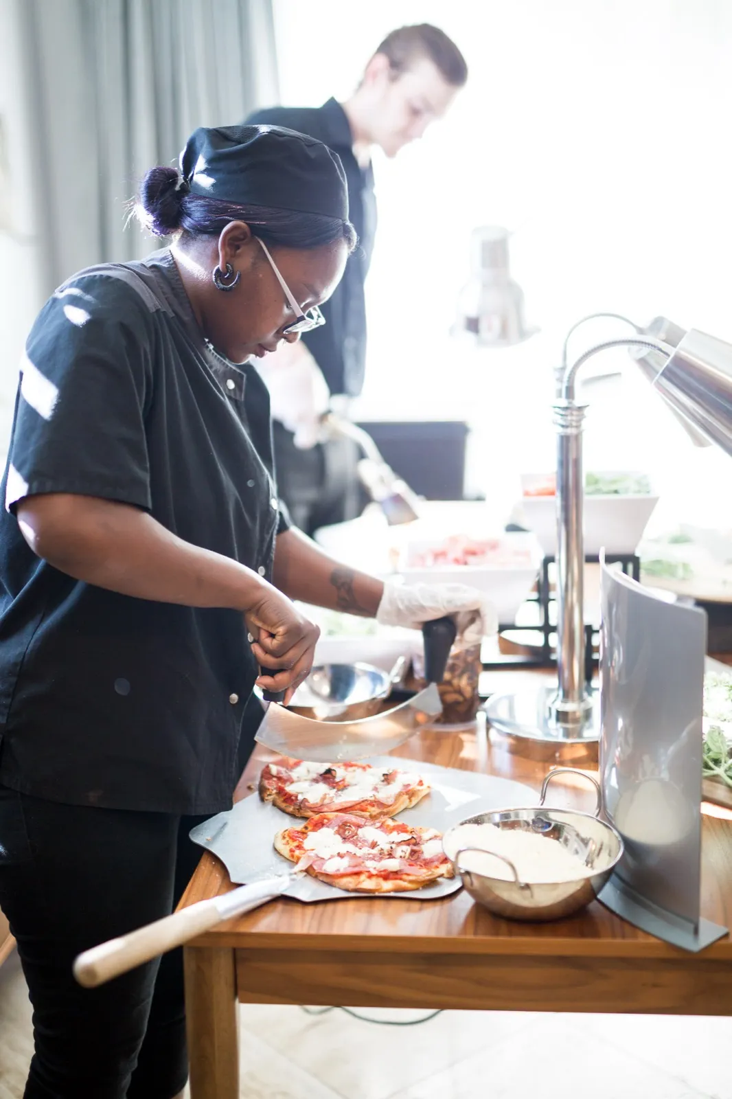 A caterer in chef’s blacks slices flatbreads at a buffet station while a second staffer works behind her