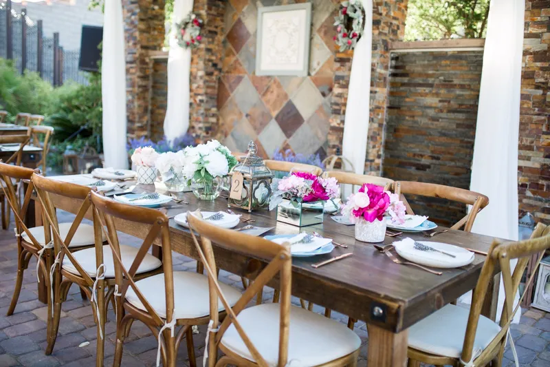 Farm tables under the pergola dressed with pink and white florals and lanterns, cross back chairs pulled up on both sides