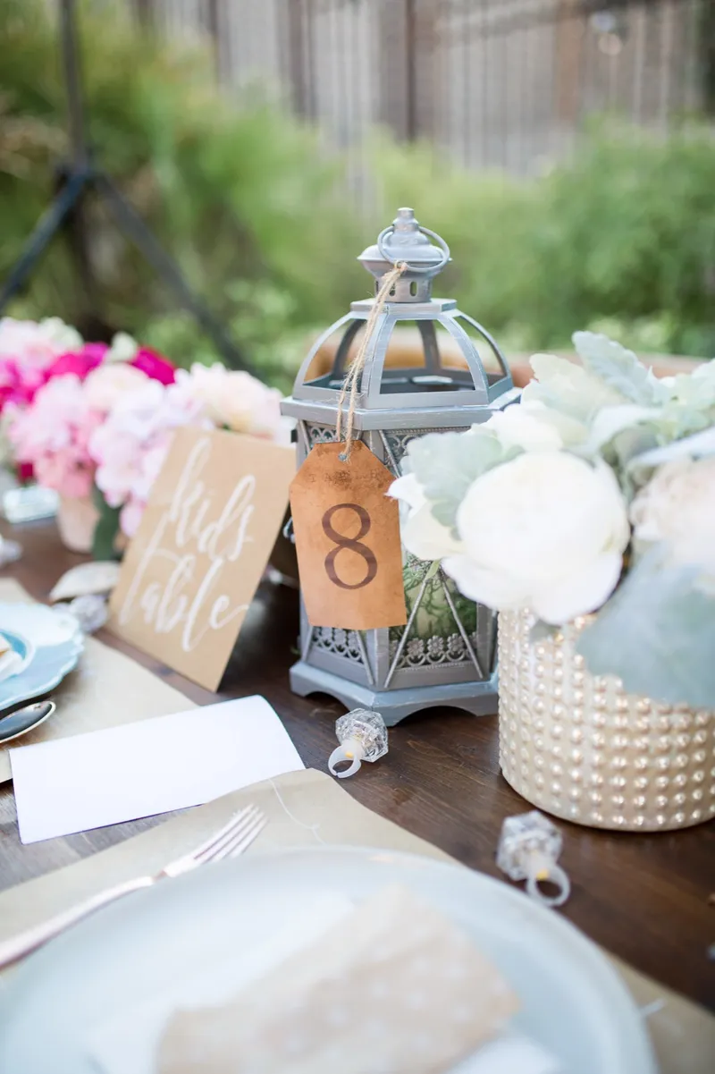 Kids table sign leaning against a lantern next to a pearl vase of white roses