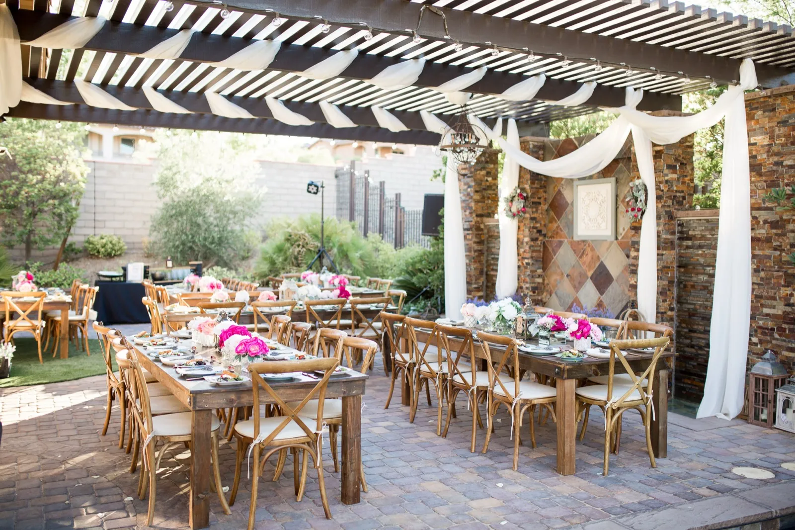Wooden farm tables and cross back chairs set for dinner under a draped pergola, with a block wall behind