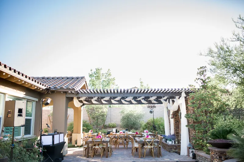 The backyard reception seen from the house side, the DJ speakers and booth on the patio, string lights along the tile roof and the tables under the pergola beyond