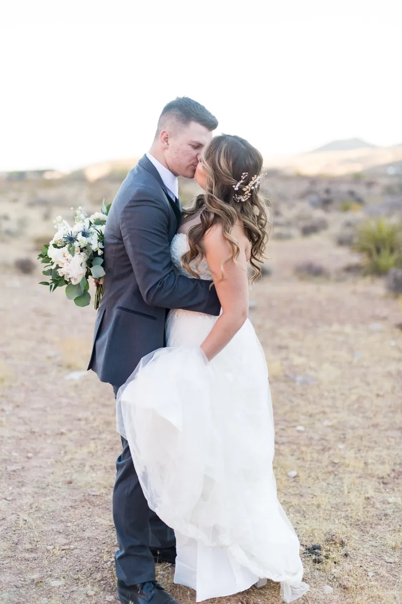 Groom kissing his bride as she holds a white bouquet, her tulle skirt catching the light