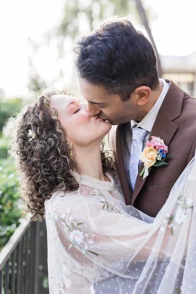 Couple kissing under the hanging willow branches, the groom in a brown suit with a peach and blue boutonniere