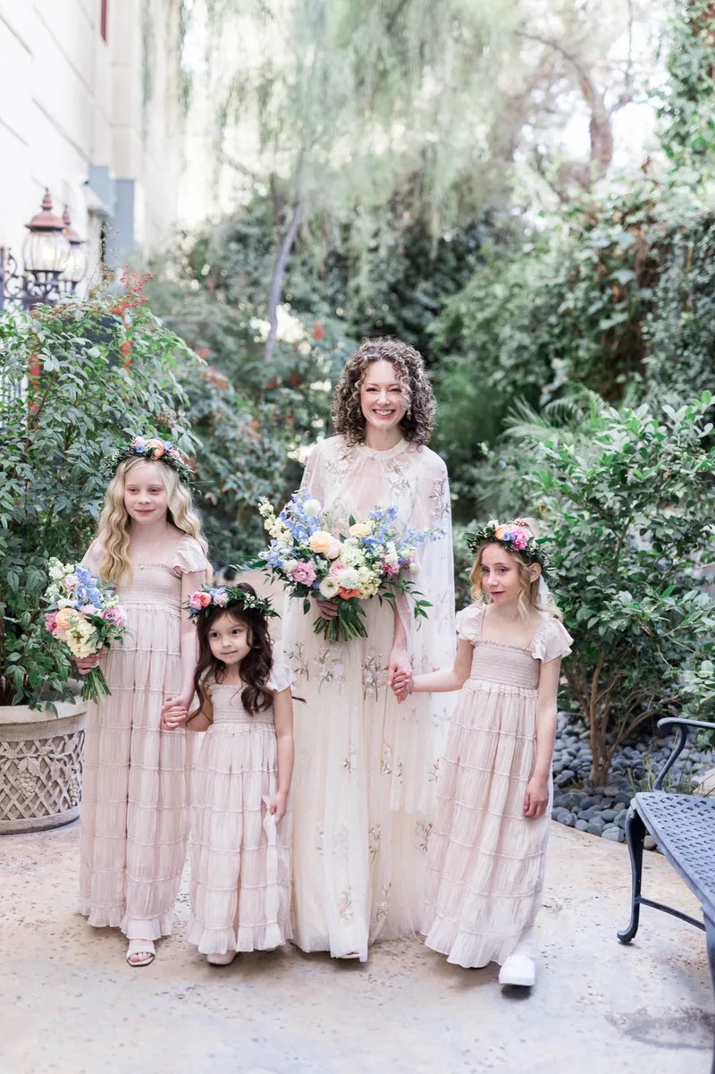 Bride standing on the garden patio with three flower girls in blush dresses and flower crowns, all of them holding bouquets