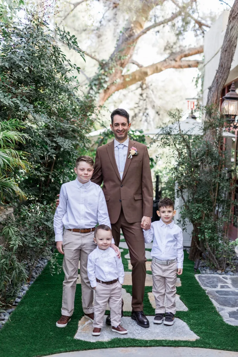 Groom in a brown suit standing on the aisle with four boys in white shirts and khakis, trees arching overhead