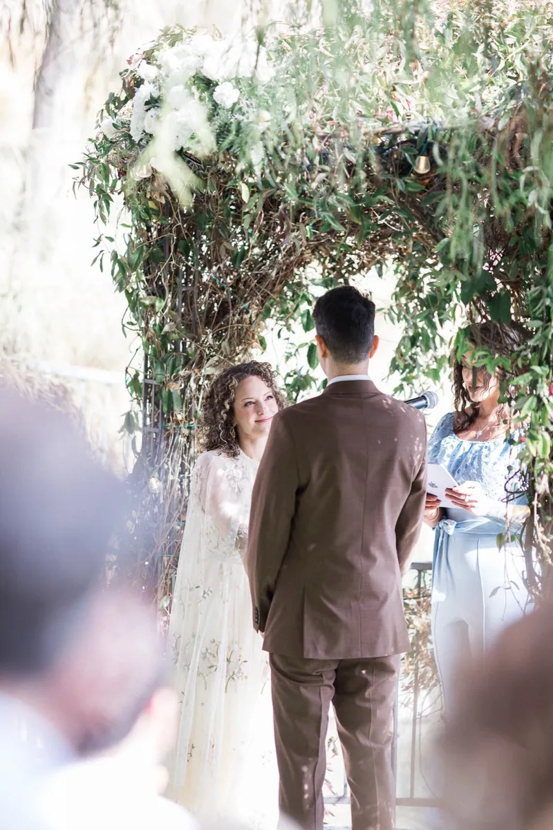 Couple facing each other at the flower topped arch with the officiant beside them and guests blurred in the foreground
