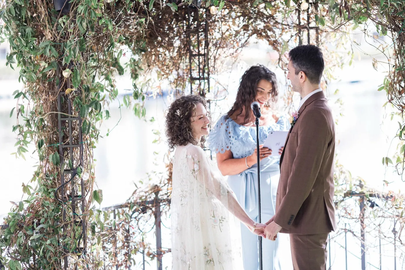 A friend in a blue dress reads from a folder at the microphone as a bride and groom hold hands under an ivy arch