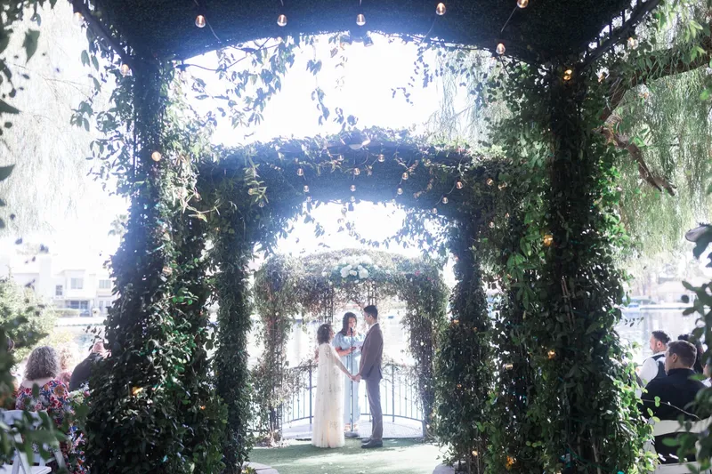 The ceremony seen from the back of the aisle, framed down the whole tunnel of arches with string lights glowing overhead