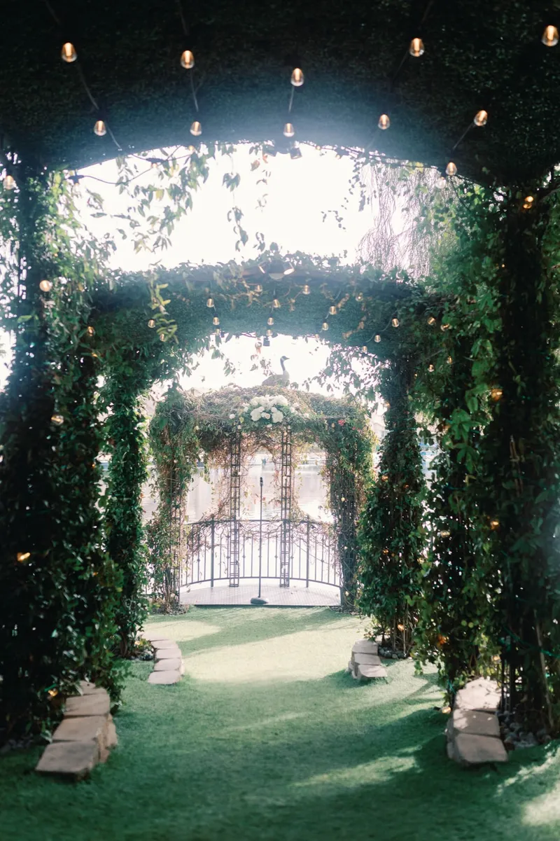 The empty aisle running under a tunnel of greenery covered arches strung with small lights, an iron gate at the far end