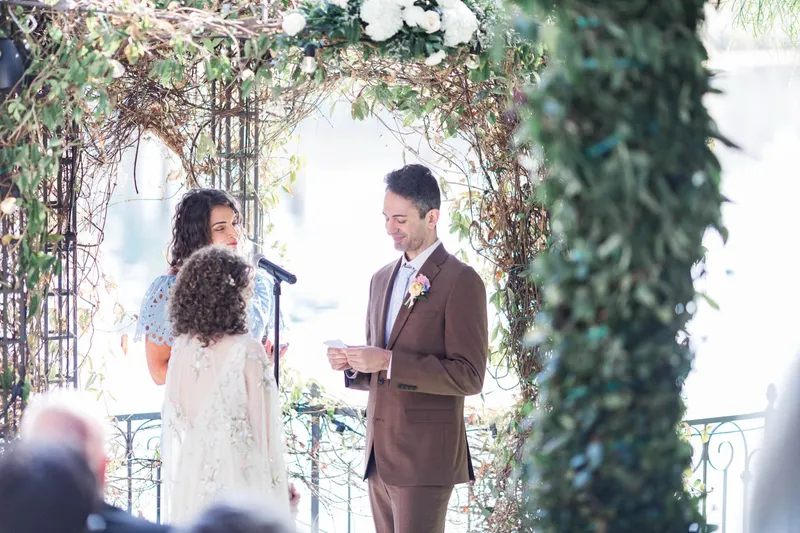 Groom reading his vows off a card under the leafy arch while the bride and the officiant listen