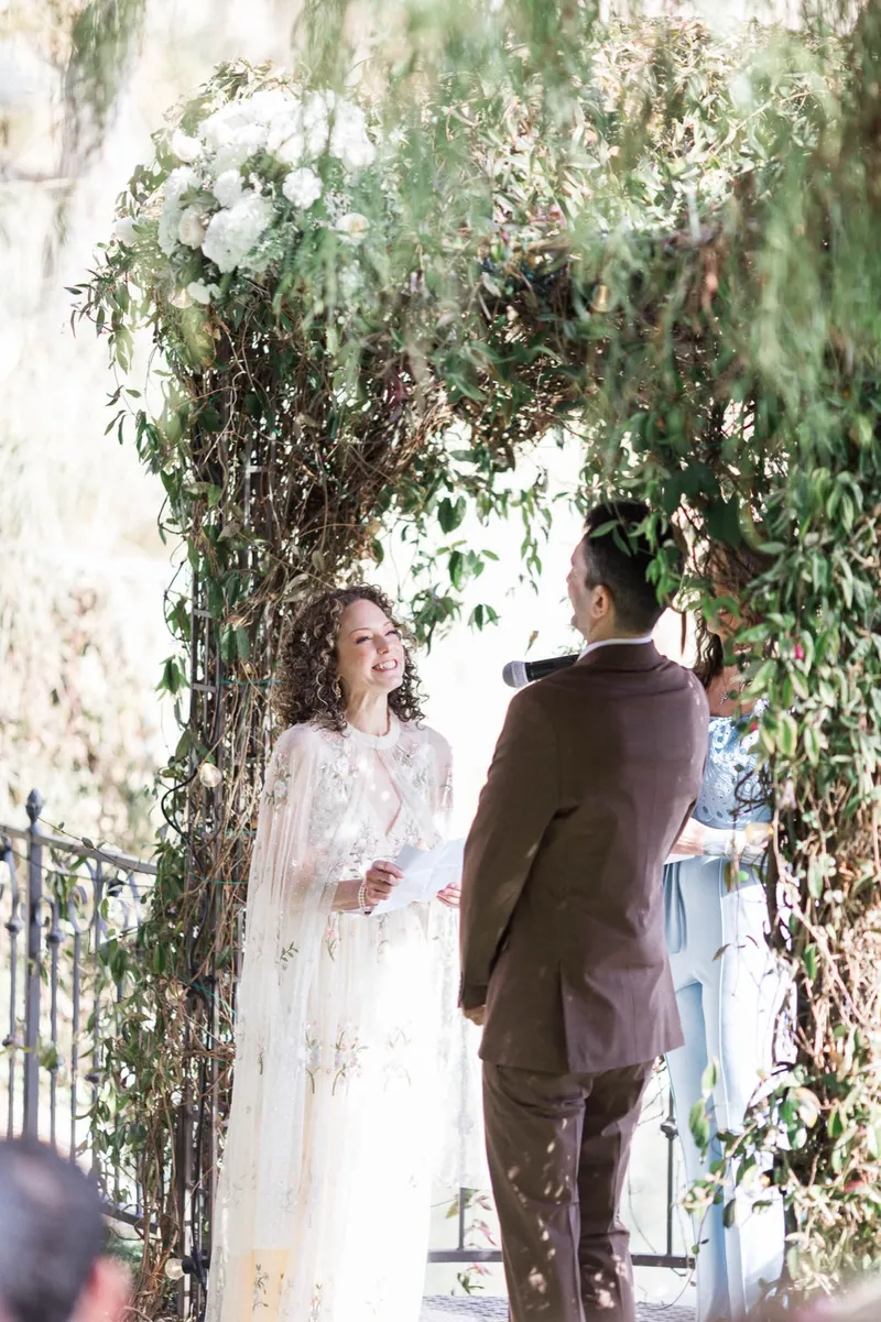 Couple standing face to face through the vows, the greenery of the arch closing in on both sides of them