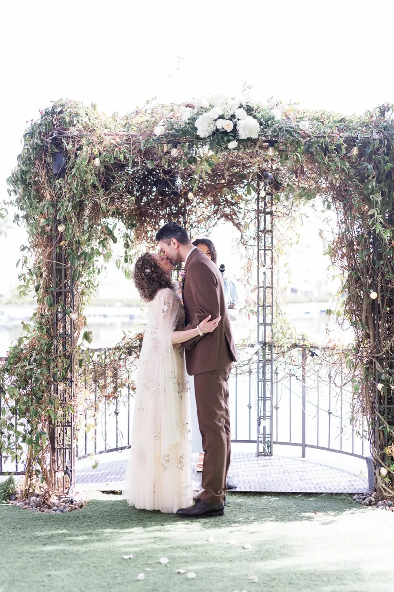 First kiss under the arch with the flower topped frame above them and the water showing through the leaves