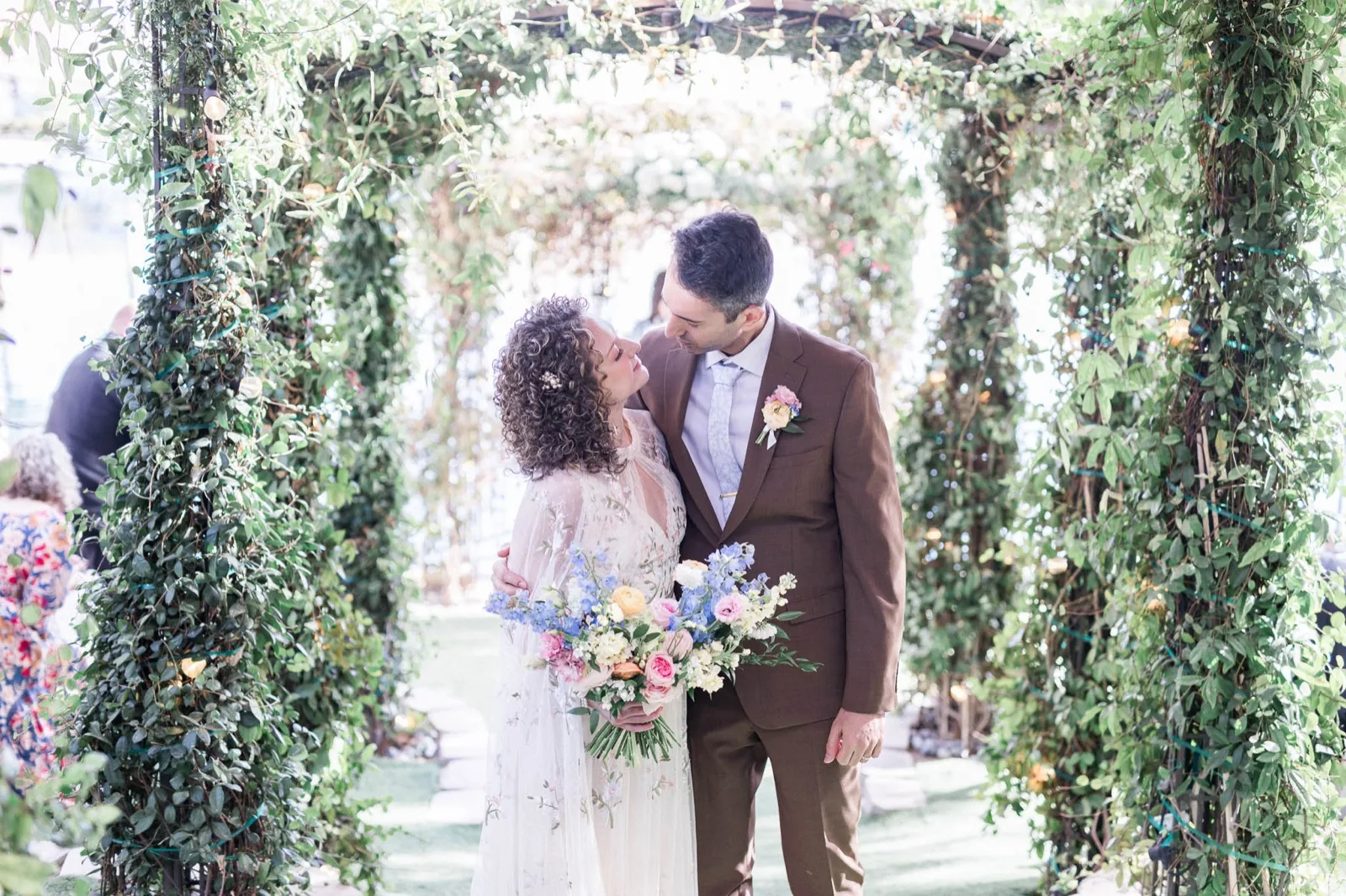Couple standing together under the greenery covered arches on the ceremony lawn, the bride holding a pastel bouquet and the lake showing through the leaves behind them