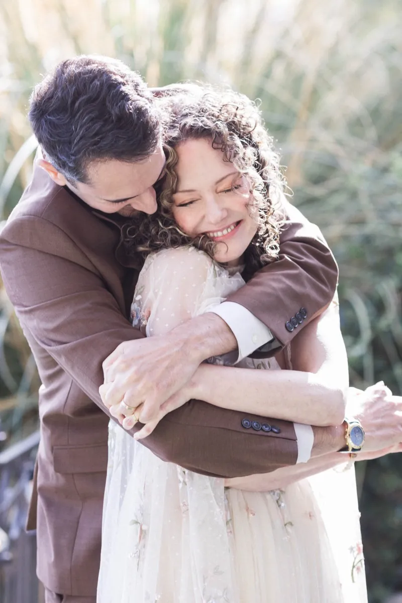 Couple holding each other tight with their eyes closed, tall ornamental grasses catching the light behind them