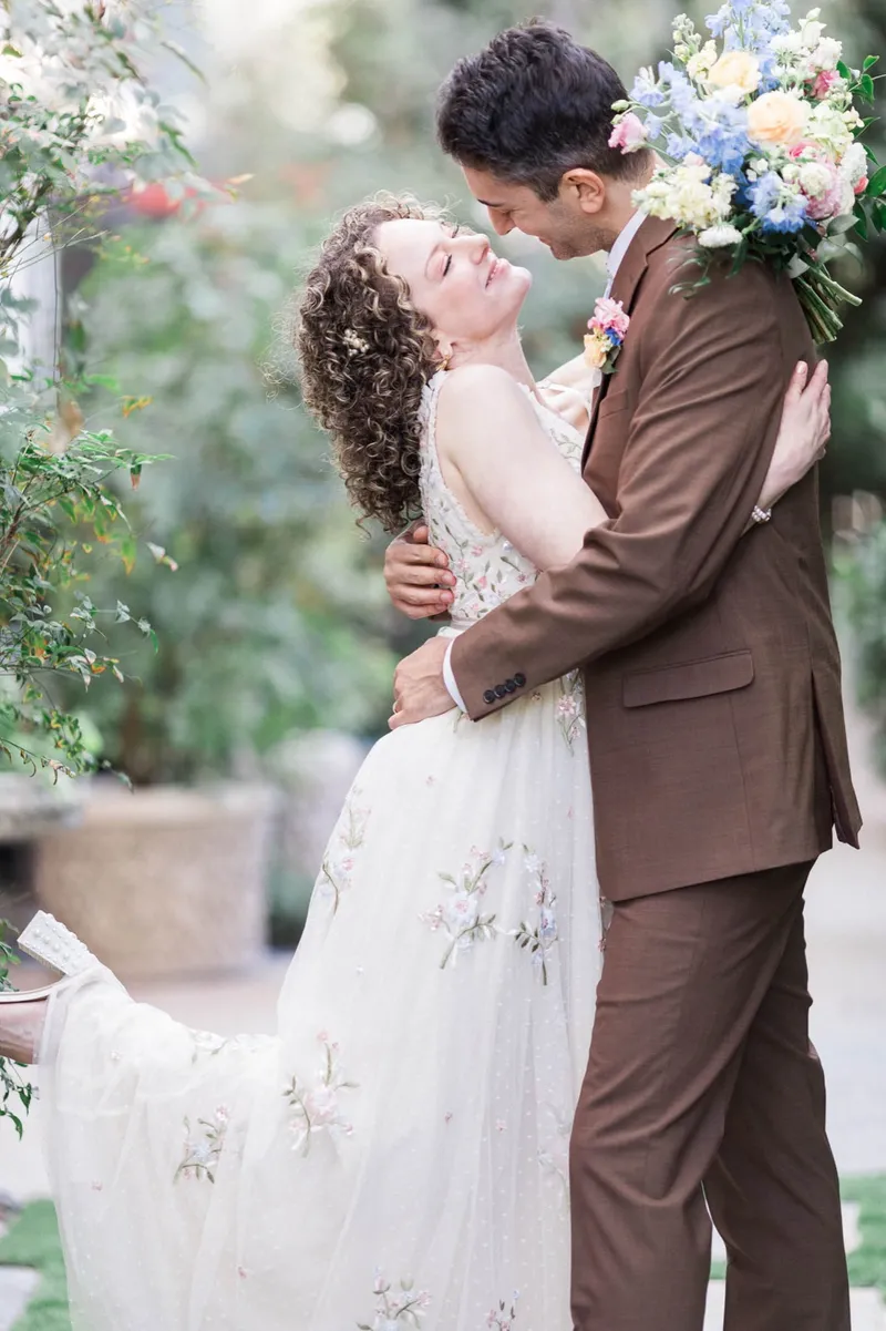 Couple leaning into a kiss with the bouquet raised at the groom's shoulder and greenery filling the background