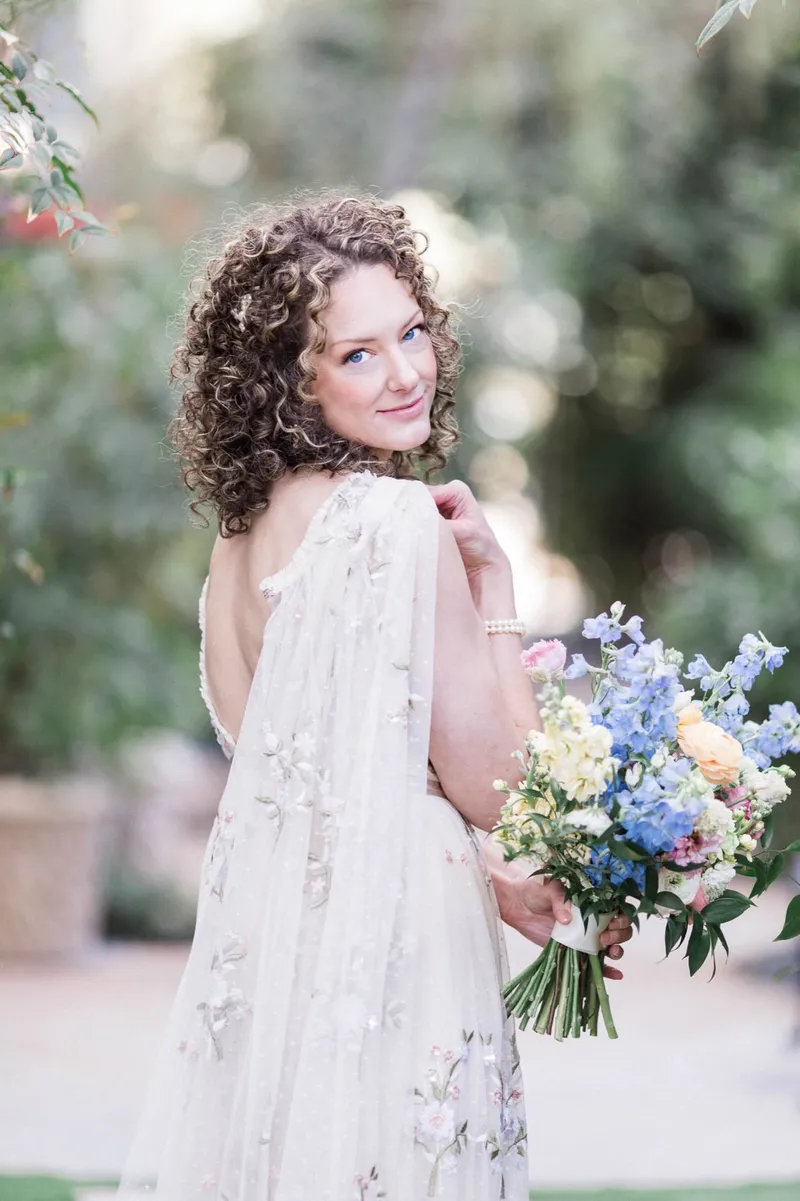 Bride looking back over her shoulder in her sheer embroidered cape, holding the pastel bouquet at her side